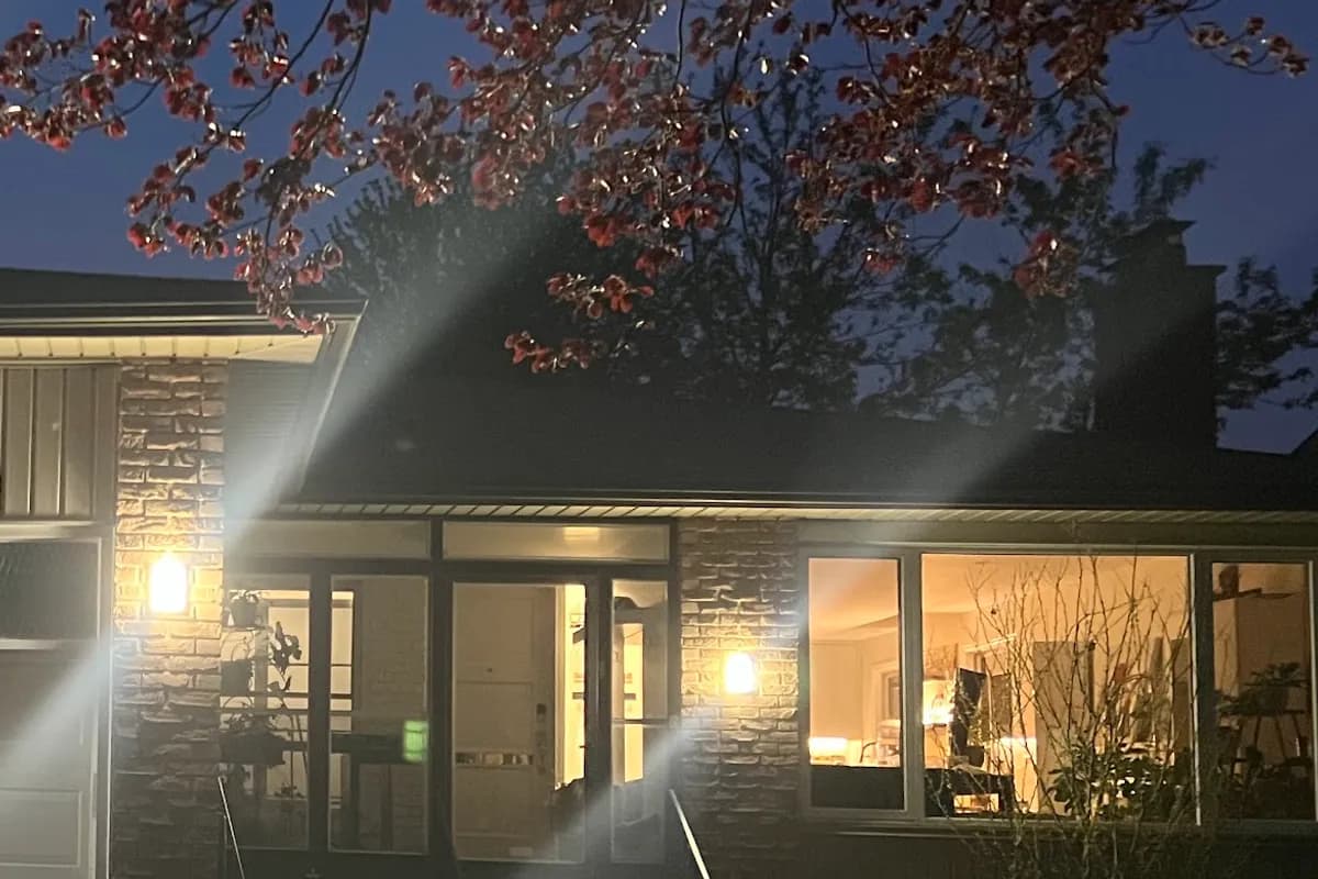 The front entrance of a brick bungalow at dusk, a wall lantern lit on each side of the glazed porch and a wide front window glowing, a flowering branch across the top of the frame.