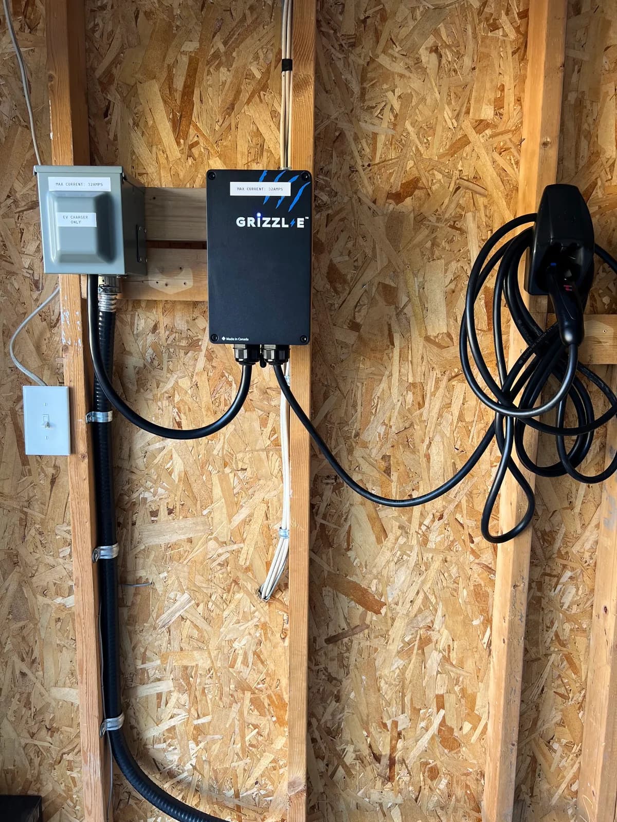 A black Grizzl-E charger on a bare OSB garage wall, a grey disconnect switch labelled “EV charger only” to its left, flexible conduit clipped down the stud and the charging cable coiled on a holster to the right.